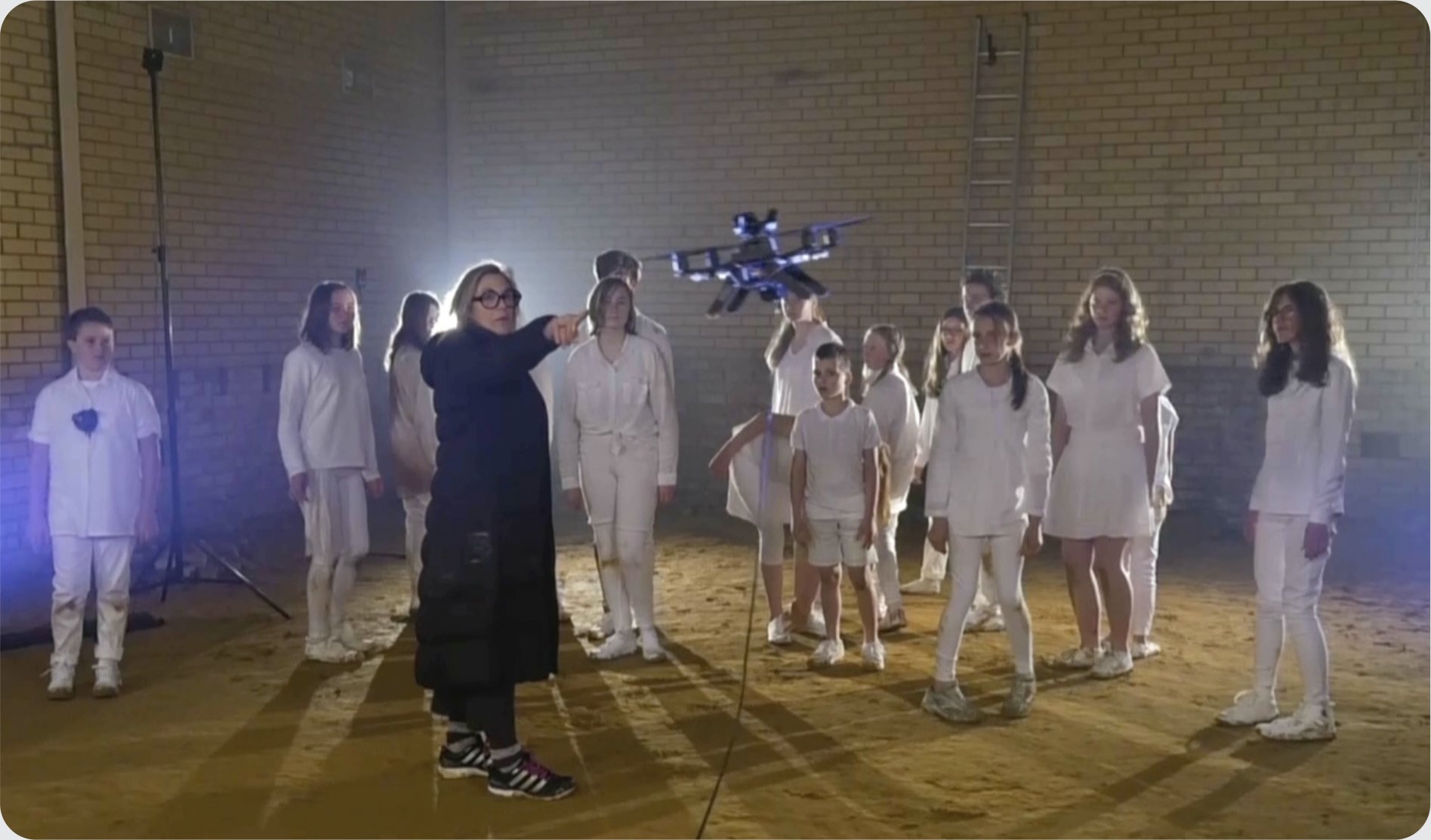 Children in white standing in a brick industrial hall while a drone hovers above them