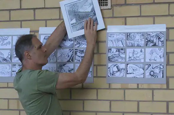 An artist pins drawings of factory buildings to a brick wall